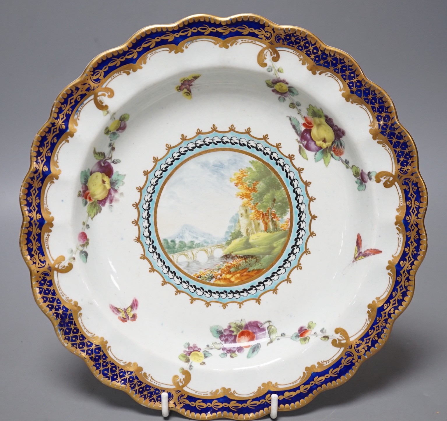 A Worcester plate painted with a landscape surrounded by fruit and moths under a blue and gilt border, in one of the variations of the Lord Dalhousie pattern (over restored chip to reverse only), 21.5 cm